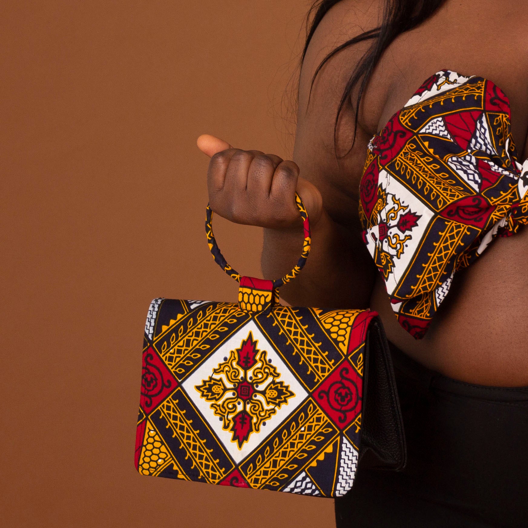 Black leather, velvet lined clutch bag overlaid with red, yellow, white and black ankara wax print in a geometric pattern with a ring handle