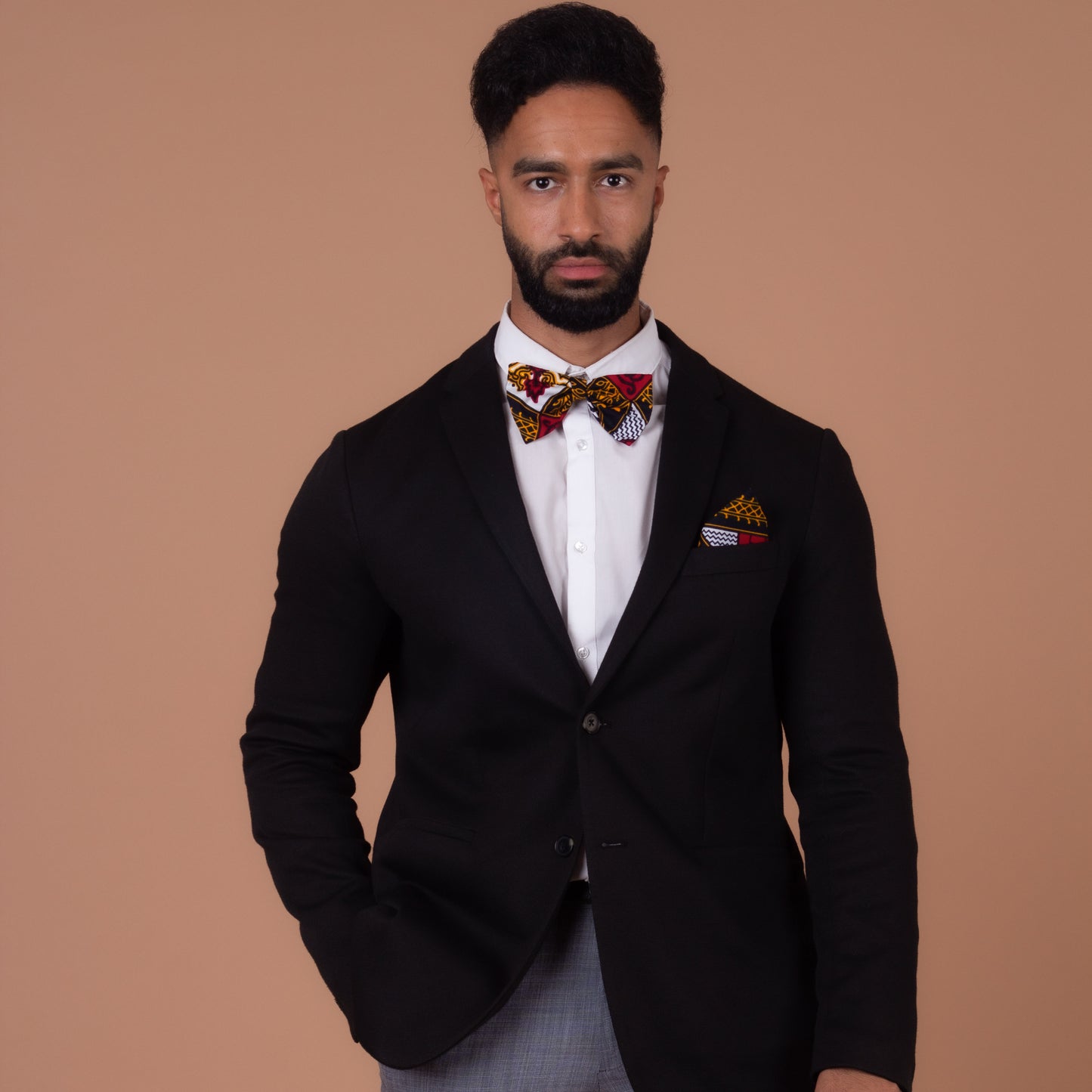 Man wearing Red, white, navy blue and yellow pocket square and bow tie with a black blazer , grey trousers and a white shirt looking directly at the camera