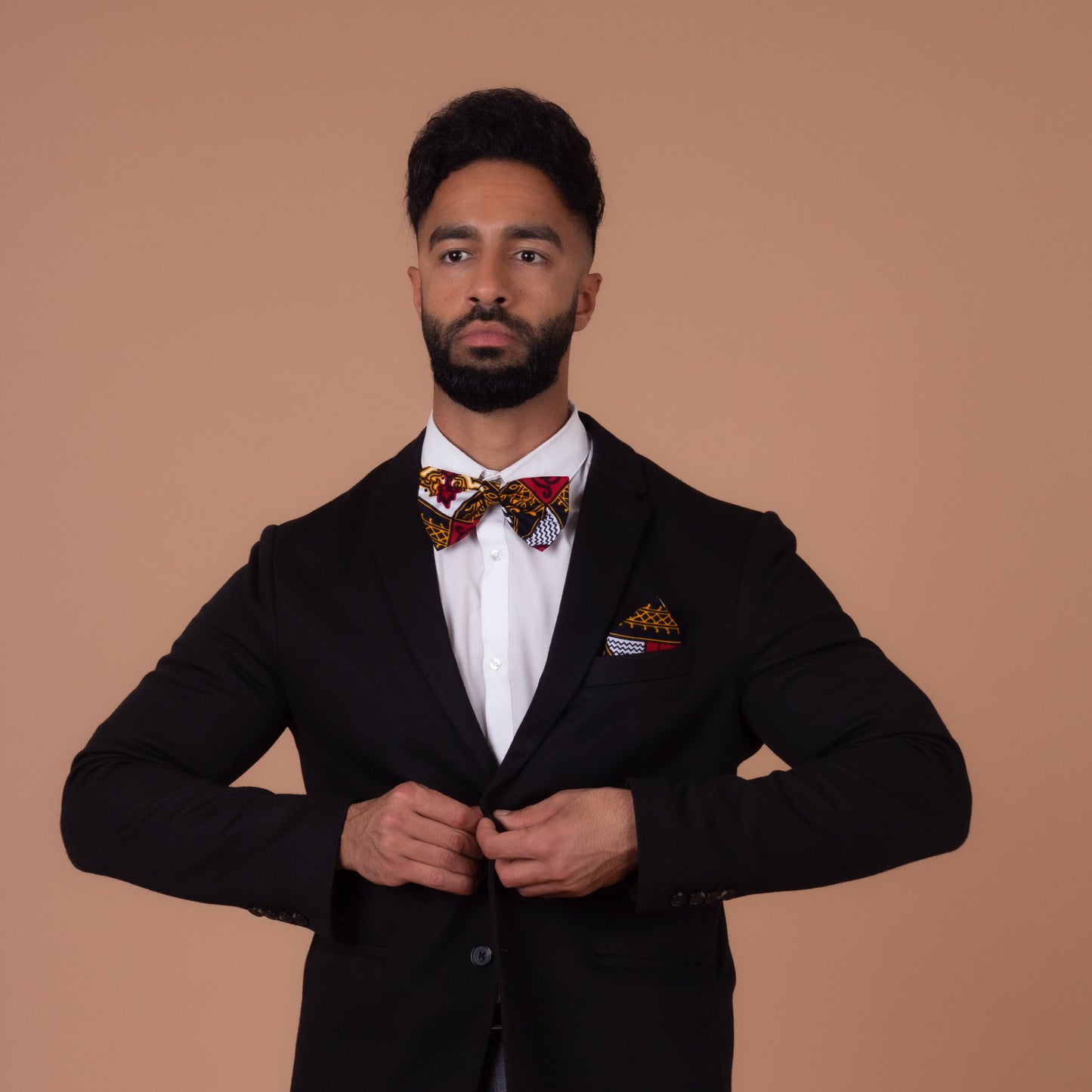 Man wearing a Red, white, navy blue and yellow pocket square and bow tie wearing a white shirt and buttoning a black blazer looking straight ahead