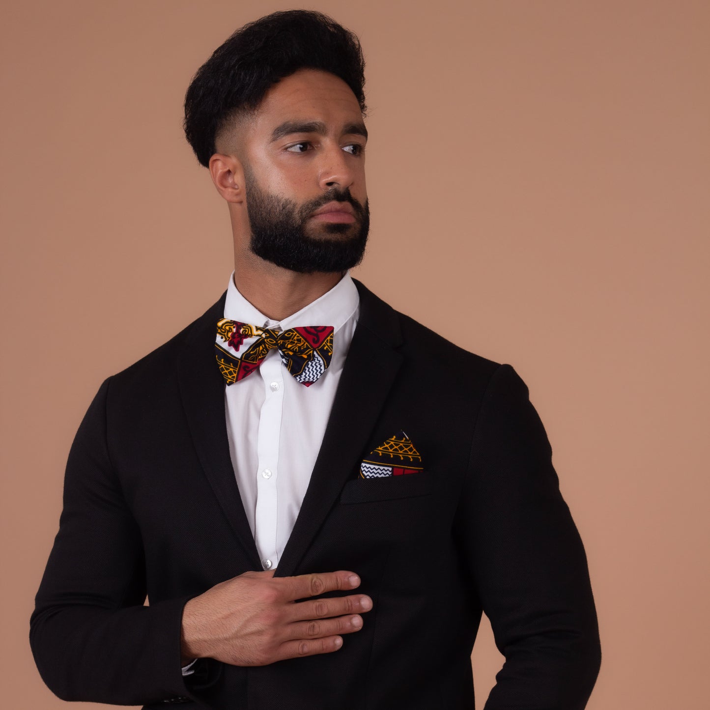 Man wearing Red, white, navy blue and yellow pocket square and bow tie with a black blazer and a white shirt looking to the left